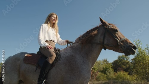 Female horsewoman enjoying active weekend day outdoors. Close-up shot of a cheerful, smiling jockey riding on a horseback with clear blue skyline in the background. High quality 4k footage