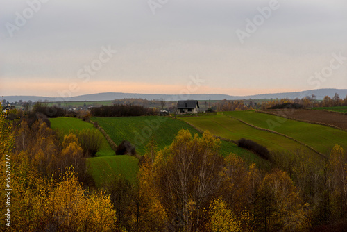 Przepiękny jesienny krajobraz . Góry Świętokrzyskie . Malownicze pola i drzewa z jesiennie wybarwionymi liśćmi ( w odcieniach pomarańczy , żółci i czerwieni ), widoczne domy i zabudowania gospodarskie