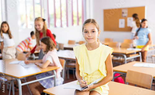 Happy tween schoolgirl repeating educational material before lesson, sitting during break on table with notebook in hands photo