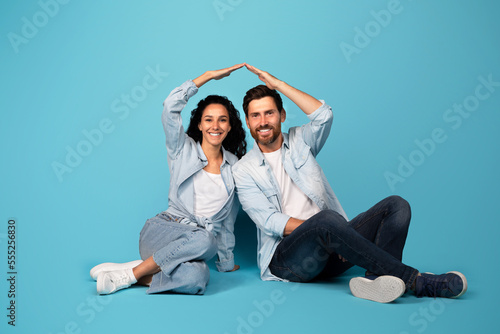 Cheerful young european couple in casual make roof sign with hands, sit on floor, isolated on blue background