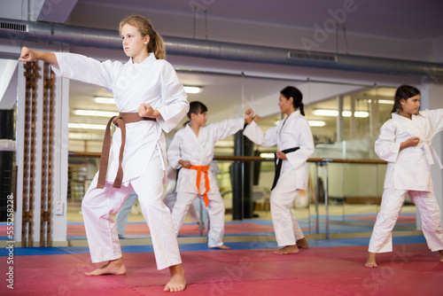 Young girl in white kimono standing in fighting stance during group karate training in gym.