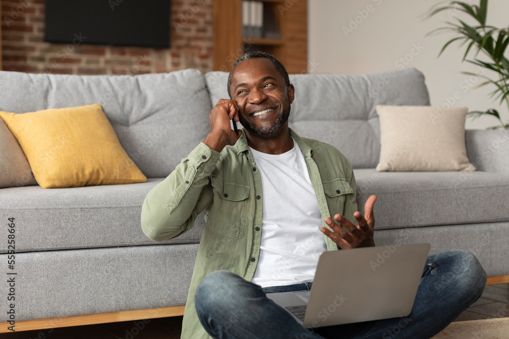 Glad adult black man uses computer, calls by phone, gesticulates, sits on floor in living room interior