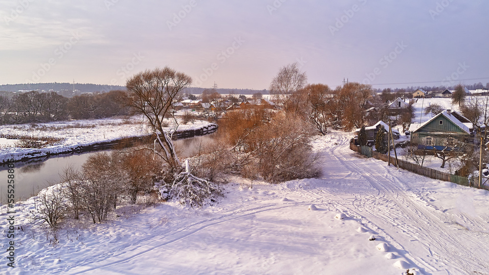 Winter river landscape, snow covered fields. Hoarfrost on trees, plants. Frosty evening. Sunny cloudy Misty weather. Cold season. Calm nature scene. Village