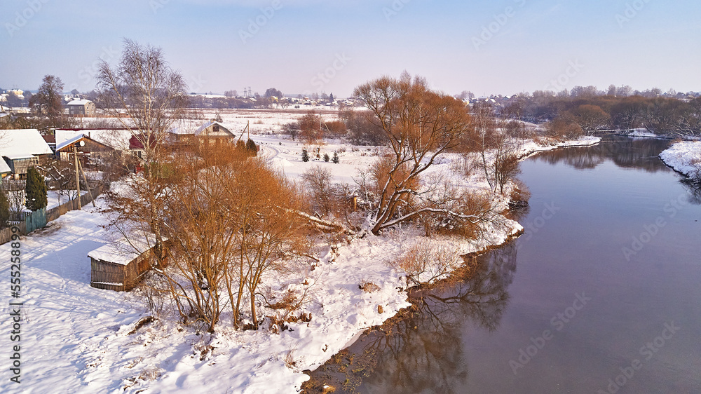 Winter river landscape, snow covered fields. Hoarfrost on trees, plants. Frosty evening. Sunny cloudy Misty weather. Cold season. Calm nature scene. Village