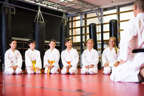 Boys and girls in white kimono kneeling on gym floor. Their karate trainer kneeling in front of them.
