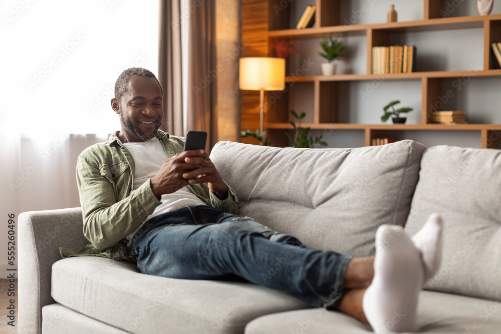 Smiling adult black guy using phone, chatting in social media, sitting on sofa in living room interior