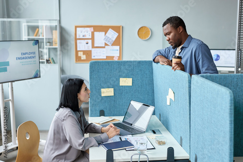 Фотография Side view at two young colleagues talking over cubicle wall in office, copy spac