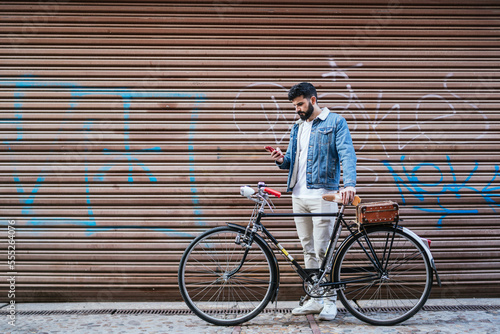 Young man in denim jacket holding his vintage classic bicycle while looking his mobile phone in front of the closing shutter of a shop.