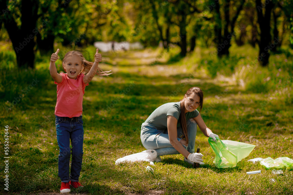 Fototapeta premium Blonde girl with eco symbol and her mom gathering garbage in the park