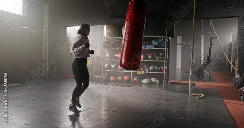 Side-view beautiful fit Arab woman professional kickboxer fighter training with punching bag in dark gym. Attractive thin energetic muslim female working out practising boxing kicking wearing hijab. photo