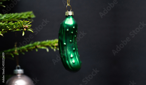 Christmas tree decorations. A green glass cucumber is hanging on a christmas tree. Green branches and a black background are visible.
