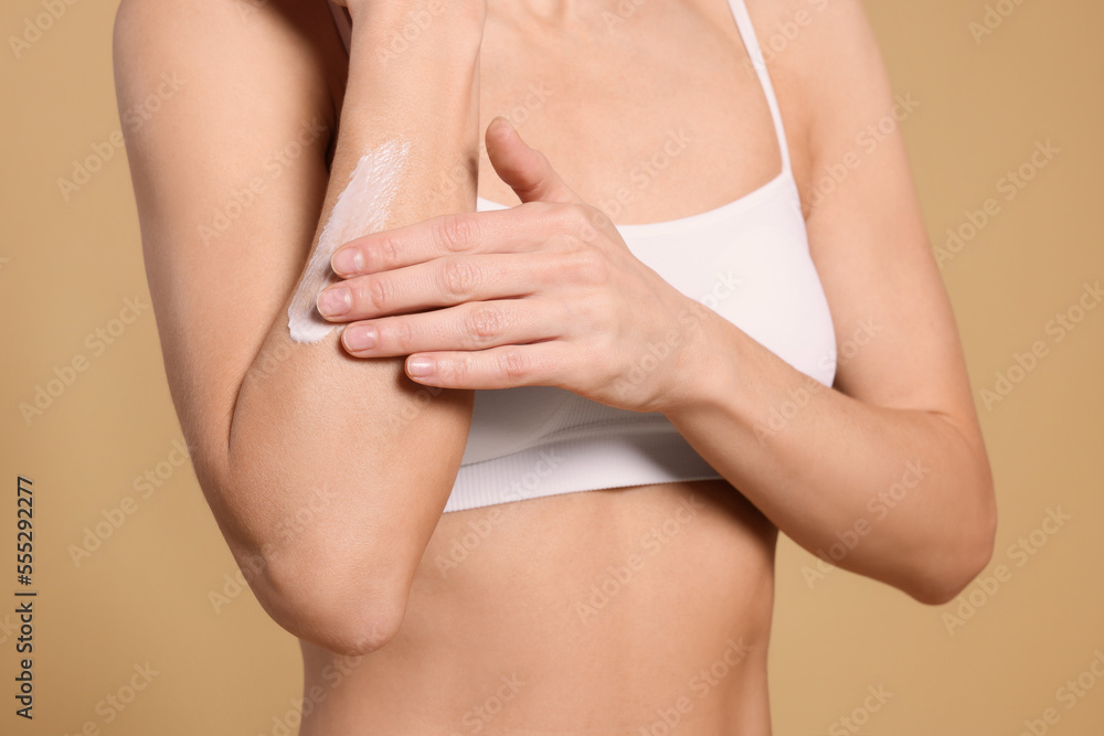 Woman applying body cream onto her arm against beige background, closeup