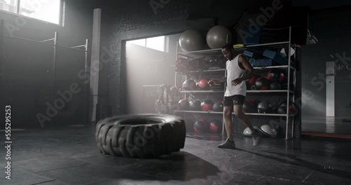 Side-view of sporty muscular African athlete lifting training with tire for crossfit in dark gym. Powerful motivated serious African-American man working out in sports clothing doing exercises.