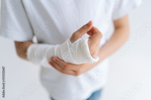 Close-up high-angle view of unrecognizable little girl with broken hand wrapped in plaster bandage massaging injured forearm on white isolated background. Concept of child insurance and healthcare.