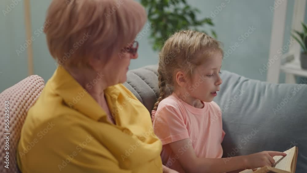 Adorable girl reads story and looks at senior woman seeking approval smiling in living room. Home education and leisure. Granny takes care of child Adorable girl reads story and looks at senior woman seeking approval smiling in living room. Home education and leisure. Granny takes care of child