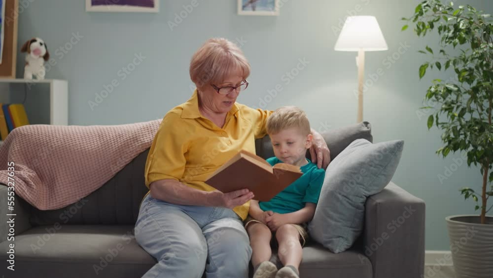 Old woman in glasses reads fairy tales to little boy resting at home slow motion. Introducing interesting stories to toddler child. Family leisure lifestyle Old woman in glasses reads fairy tales to little boy resting at home slow motion. Introducing interesting stories to toddler child. Family leisure lifestyle
