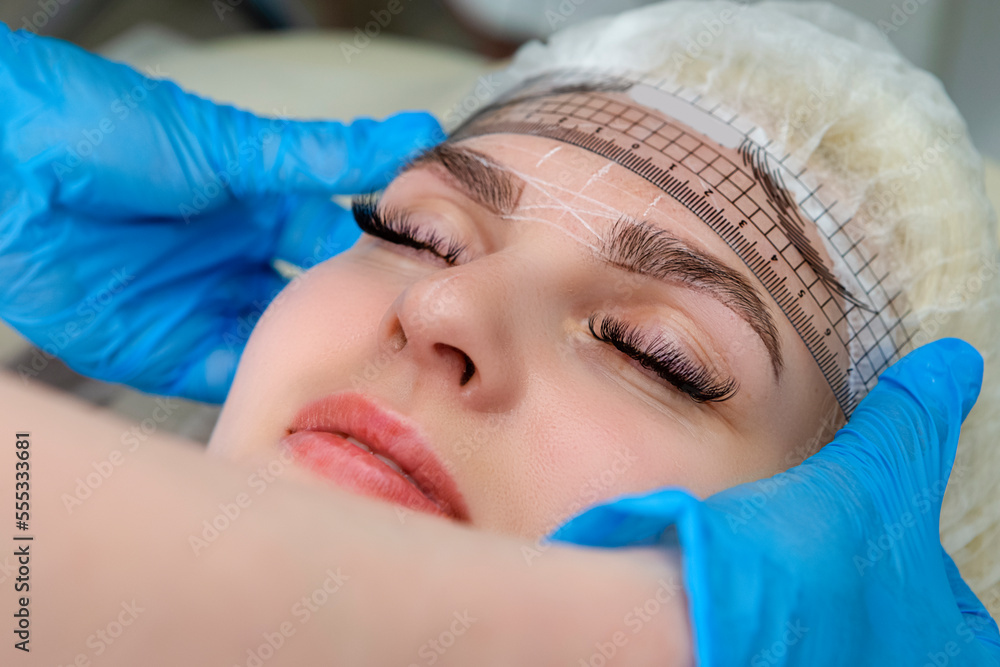 Closeup of Facial Tattoo Markup with Transparent Facial Ruler On Eyebrows of Young Caucasian Woman During a Process of Permanent make up in Salon. Closeup of Facial Tattoo Markup with Transparent Facial Ruler On Eyebrows of Young Caucasian Woman During a Process of Permanent make up in Salon.