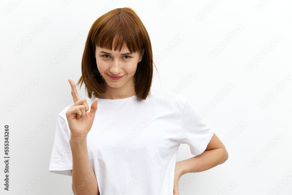 a pleasant, beautiful, attractive woman stands on a white background in a white T-shirt and looking at the camera shows her index finger up