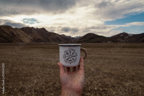 Close up person holding enamel mug concept photo. Drinking at hike. First view hand photography with large mountain on background. High quality picture for wallpaper, travel blog, magazine, article