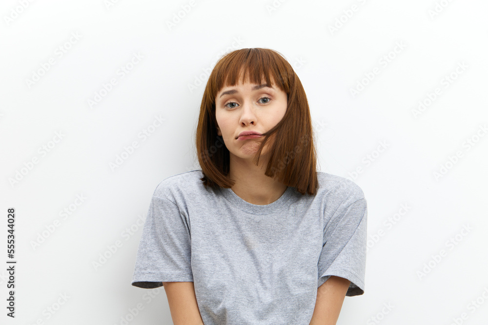 a sad, upset woman stands on a white background in a gray T-shirt and with a strand stuck to her face looks sadly at the camera