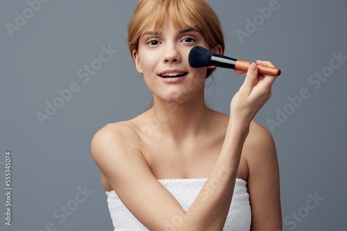 a happy, joyful woman stands on a gray background wrapped in a towel and paints her face with a fluffy makeup brush, smiling pleasantly