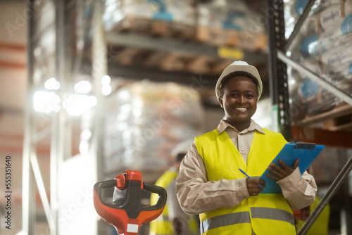 A smiling African-American woman is working in a distribution warehouse taking inventory and moving packages.
