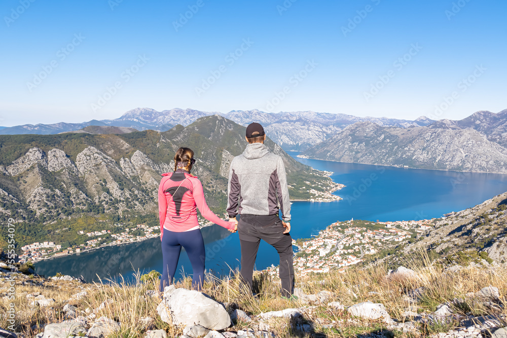 Loving couple holding hand with panoramic view of Kotor bay in sunny summer, Adriatic Mediterranean Sea, Montenegro, Balkan, Europe. Fjord winding along coastal towns. Lovcen and Orjen mountain range