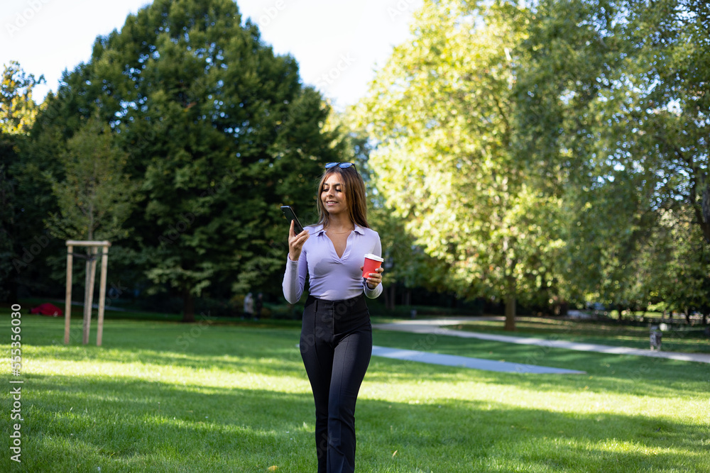Woman walking in the park and reads a message on her smartphone.  Portrait woman with brown hair, cup of coffee and mobile phone. Outdoor, public park, relaxed position and summer. 