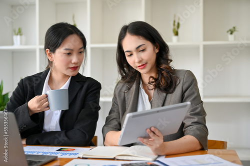 Two professional Asian businesswomen working, looking at tablet screen, discussing