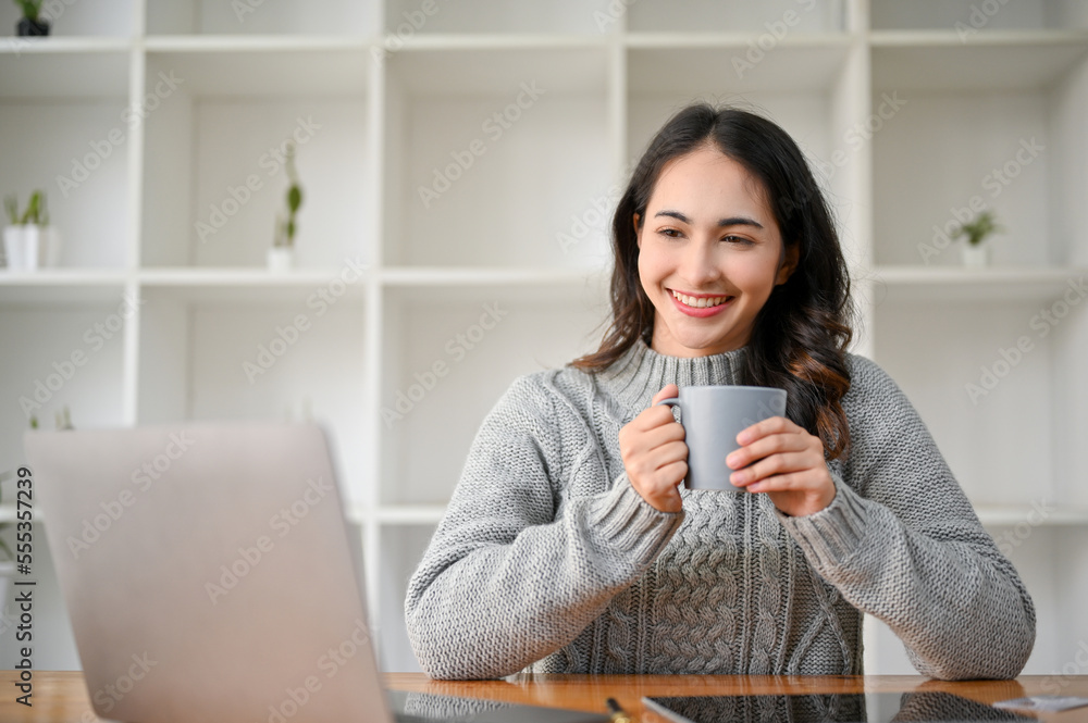 Charming Asian businesswoman looking at laptop screen, holding a coffee mug, sitting at her desk. Charming Asian businesswoman looking at laptop screen, holding a coffee mug, sitting at her desk.