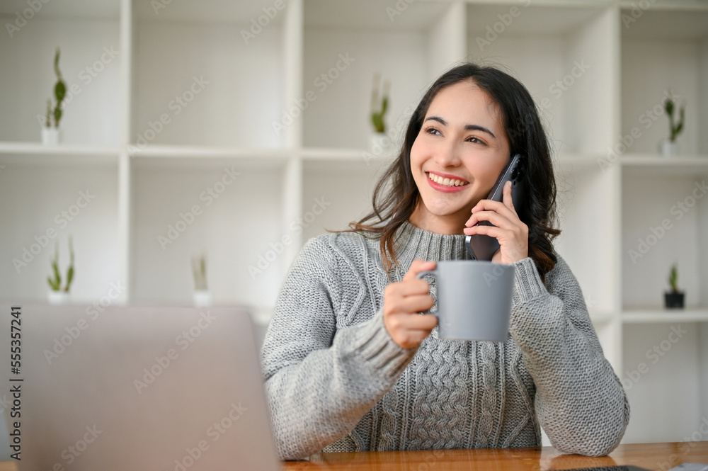 Happy Asian businesswoman sits at her desk sipping coffee while talking on the phone with someone. Happy Asian businesswoman sits at her desk sipping coffee while talking on the phone with someone.