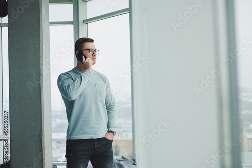 A successful man in casual clothes is standing by the window in the workspace and talking on the phone. A young manager works remotely.