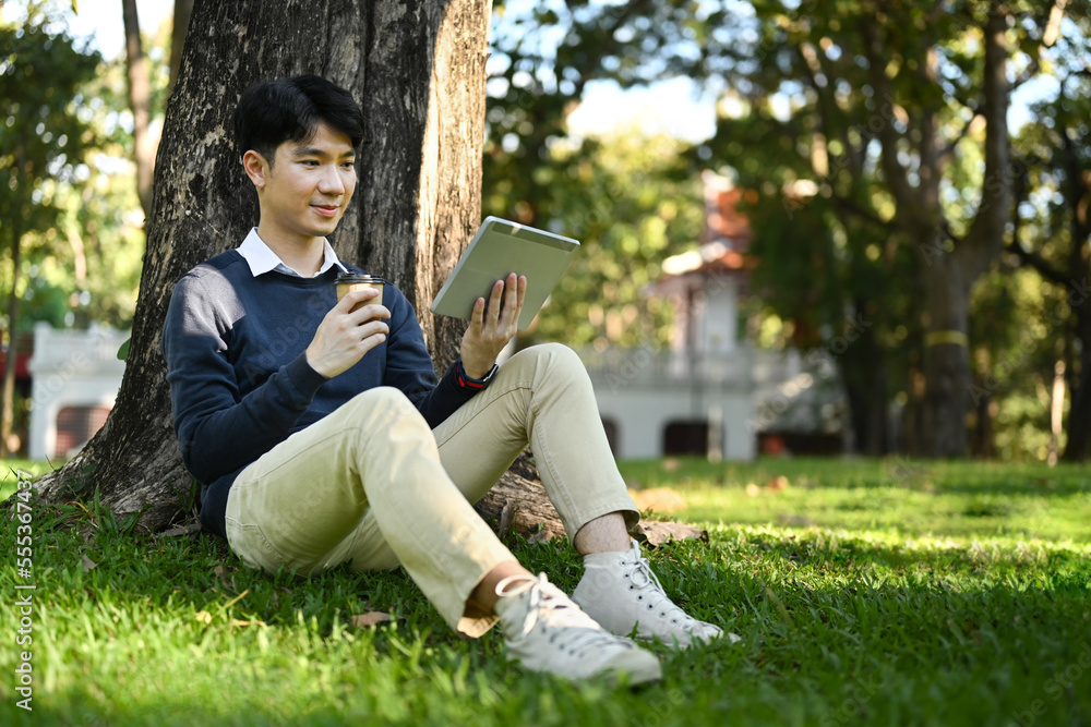 Pleased asian man drinking coffee and using digital tablet under tree on green grass at sunny beautiful day Pleased asian man drinking coffee and using digital tablet under tree on green grass at sunny beautiful day