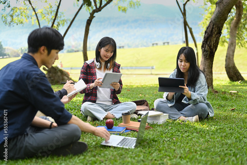 Group of university students working and reading book while sitting together on green lawn at university campus