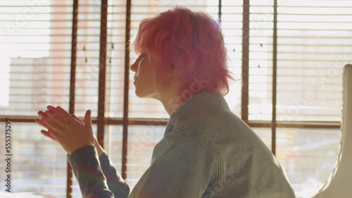 Side view slow motion portrait of apathic young gen Z woman with short pink hair wearing casual slothes sitting indoors thinking about something photo