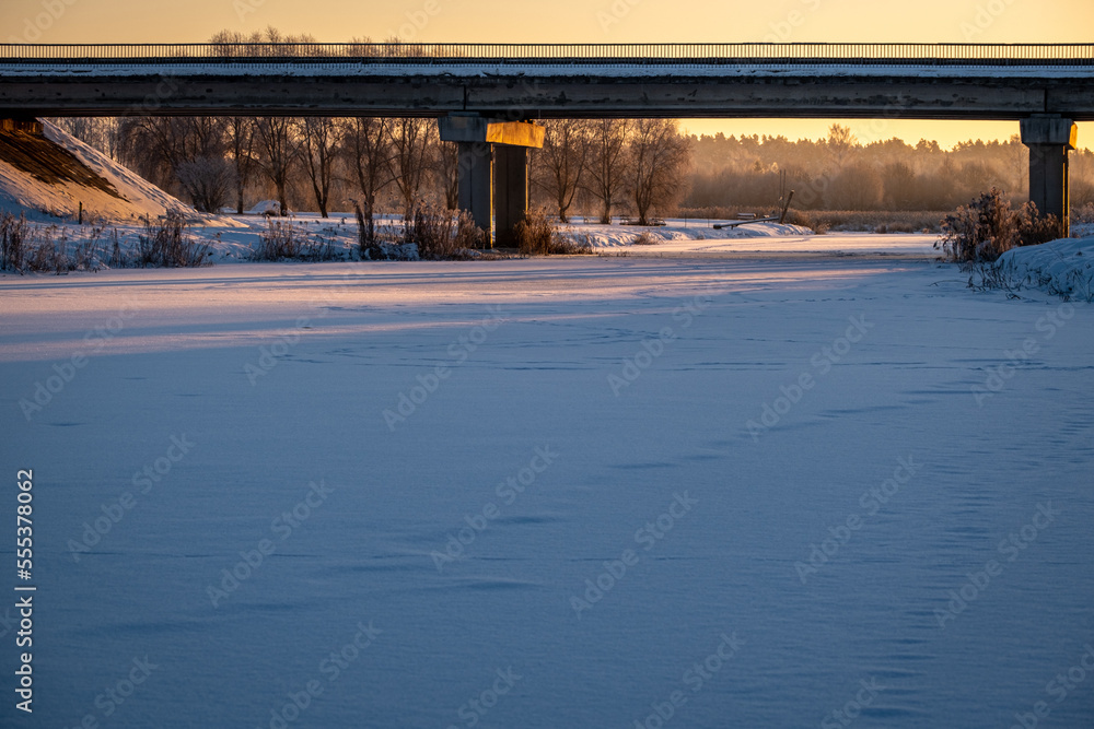 transport bridge in golden sunrise light over river Vircava near Jelgava town, Latvia. Snow covered water surface with strong safe ice cover for walking and winter fishing