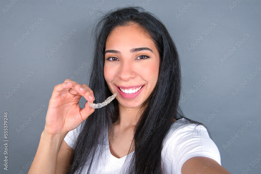 Young beautiful hispanic woman wearing white t-shirt over gray studio background, holding an invisible aligner on one hand and smiling. Young beautiful hispanic woman wearing white t-shirt over gray studio background, holding an invisible aligner on one hand and smiling.