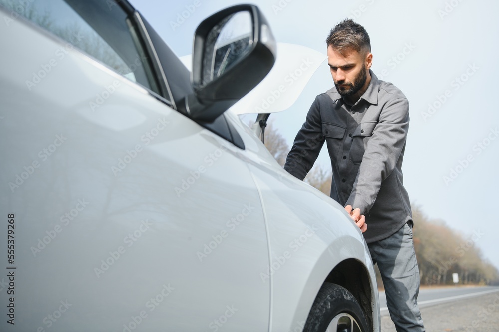A young man near a broken car with an open hood on the roadside. A young man near a broken car with an open hood on the roadside.