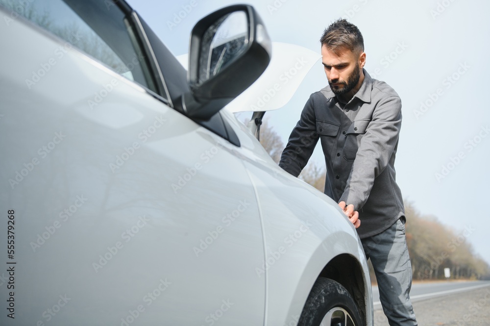 A young man near a broken car with an open hood on the roadside.