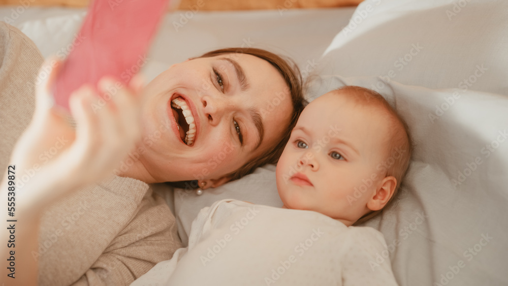 Young Beautiful Mother Lying in a Bed with Her Baby Girl, Making a Video Call on Smartphone. Having a Casual Talk with Family or Friends. Happy Female Waving into the Phone Camera. Young Beautiful Mother Lying in a Bed with Her Baby Girl, Making a Video Call on Smartphone. Having a Casual Talk with Family or Friends. Happy Female Waving into the Phone Camera.