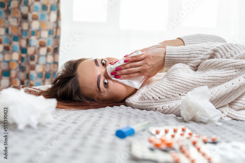 A young Caucasian woman is lying on the bed next to medicines, blowing her nose in a handkerchief. Side view. Concept of viral infection and quarantine photo