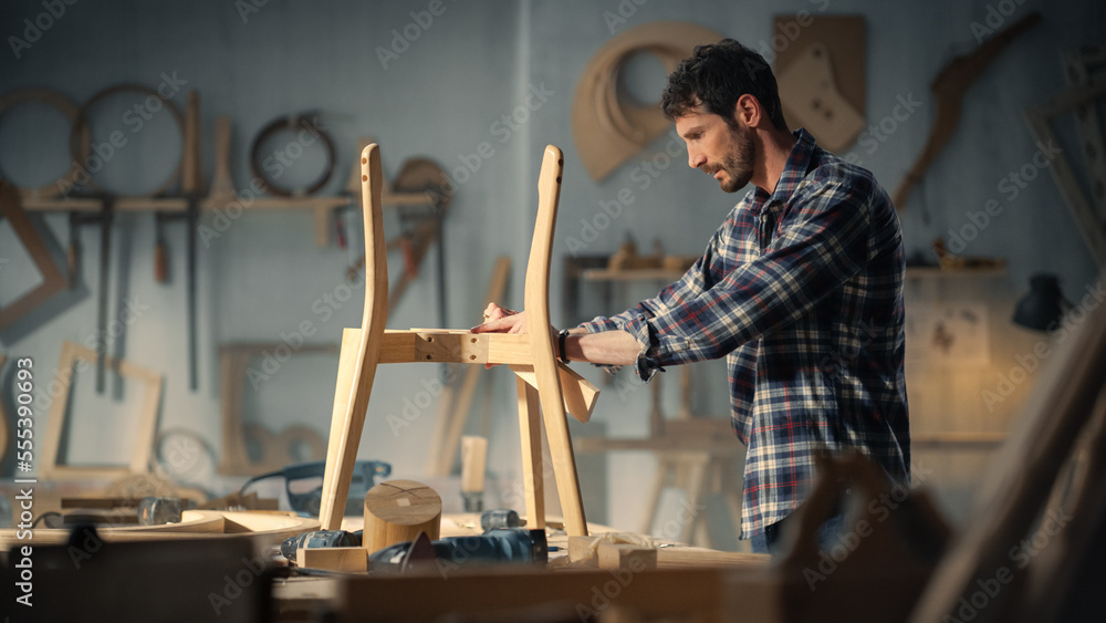 Young Carpenter Reading Blueprints and Starting to Assemble Parts of a Wooden Chair with a Rubber Hammer. Stylish Furniture Designer Working in a Studio in Loft Space with Tools on the Walls. Young Carpenter Reading Blueprints and Starting to Assemble Parts of a Wooden Chair with a Rubber Hammer. Stylish Furniture Designer Working in a Studio in Loft Space with Tools on the Walls.