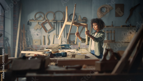 Young Female Carpenter Reading Blueprint, Making Notes, Starting to Assemble Parts of a Wooden Chair. Stylish African American Furniture Designer Working in a Studio in Loft with Tools on Walls.
