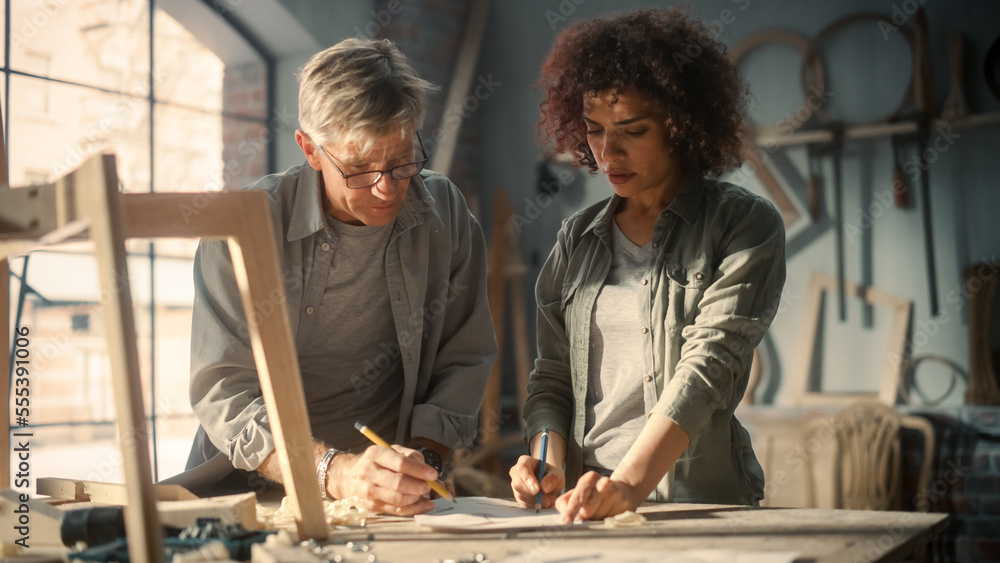 Small Business Owners of a Furniture Workshop Using Laptop Computer and Discussing the Design of a New Wooden Chair. Middle Age Carpenter and a Young Female Apprentice Working in Loft Studio. Small Business Owners of a Furniture Workshop Using Laptop Computer and Discussing the Design of a New Wooden Chair. Middle Age Carpenter and a Young Female Apprentice Working in Loft Studio.