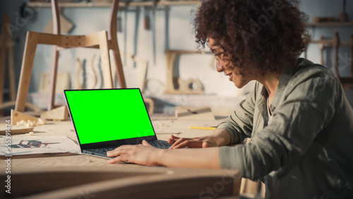 Over the Shoulder Shot of a Furniture Designer Working on Laptop Computer with Green Screen Mock Up Display. Creative Female Preparing a Design for Carpentry Project.