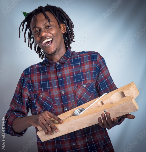 Portrait, prop and happy man holding giant peg looking cheerful on a grey studio background. Face, front and laughing, joyful african american male holding a clothes peg on a studio backdrop