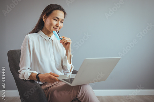 Online Shopping. Positive Young Girl Using Laptop and Credit Card At Home, Making Purchases In Internet, Sitting On a Chair In Living Room. Cybershopping concept.  photo