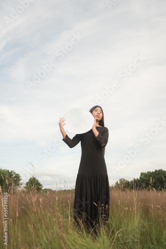 fine art portrait of happy woman in black dress standing in field holding round mirror reflecting cloudy sky photo