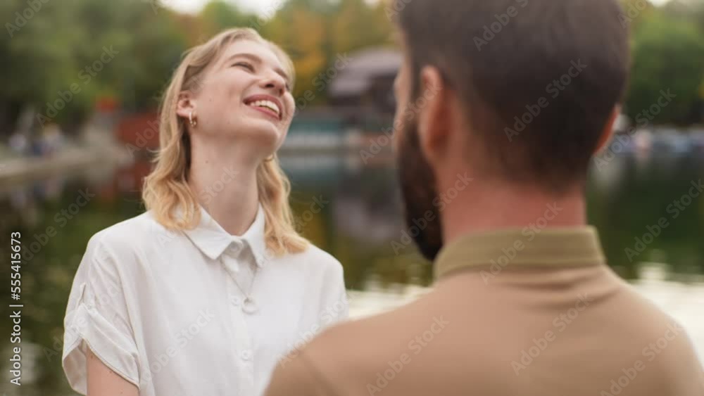 Close-up view from back of unrecognizable bearded man to happy charming man standing holding hands with boyfriend outdoors by beautiful lake at city park, talking and looking at each other smiling. Close-up view from back of unrecognizable bearded man to happy charming man standing holding hands with boyfriend outdoors by beautiful lake at city park, talking and looking at each other smiling.