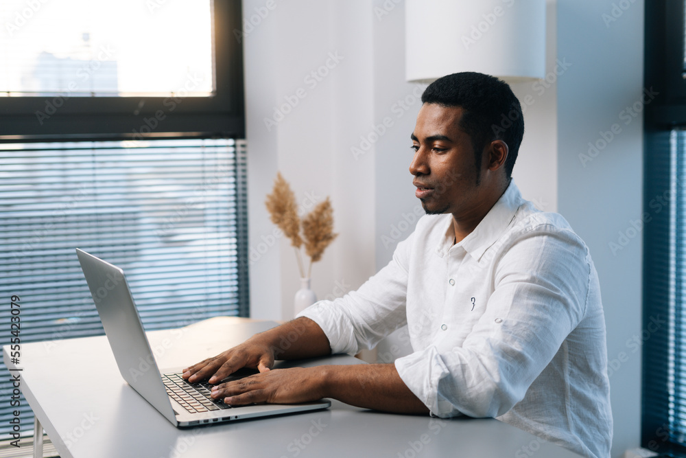 Side view of confident young African-American male entrepreneur working typing on business project on laptop sitting at desk in modern office room with light interior, on background of window. Side view of confident young African-American male entrepreneur working typing on business project on laptop sitting at desk in modern office room with light interior, on background of window.